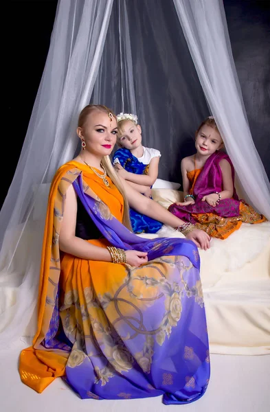 Joven rubia en un brillante saris indio y joyas sentada bajo un valance transparente con dos hijas jóvenes — Foto de Stock