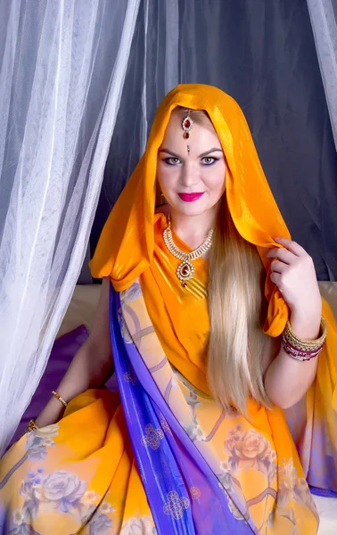 Mujer rubia joven en un brillante saris indio y joyas mirando al espectador mientras se sienta bajo un valance —  Fotos de Stock
