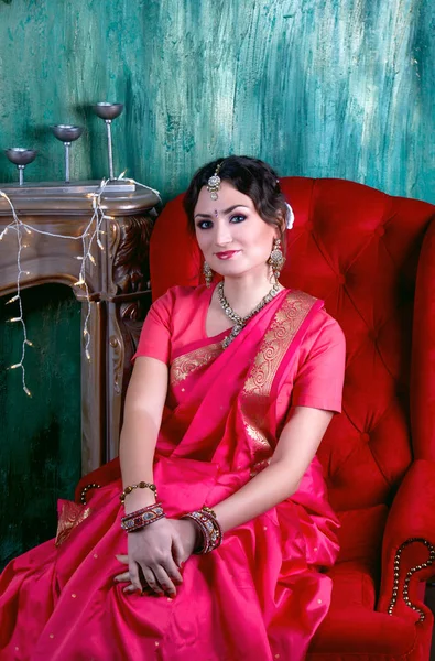 La chica en un sari escarlata y joyas indias se sienta sonriendo en el sillón rojo suave cerca de la chimenea en el fondo de la pared verde — Foto de Stock