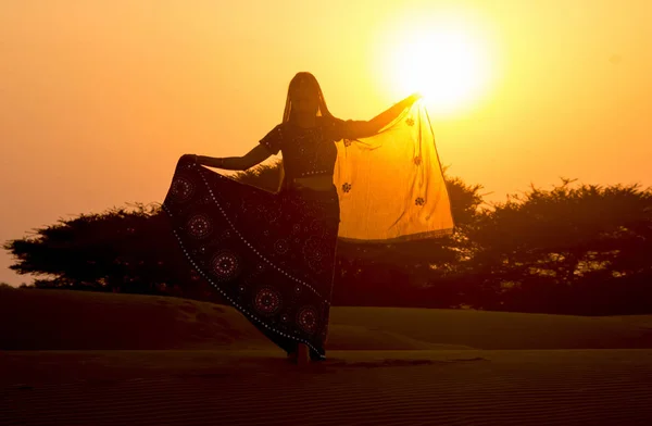 Giovane donna in abiti indiani balla al tramonto nel deserto sulle dune alzando la mano al sole contro bassi cespugli e cielo. Rajasthan, India . Immagini Stock Royalty Free