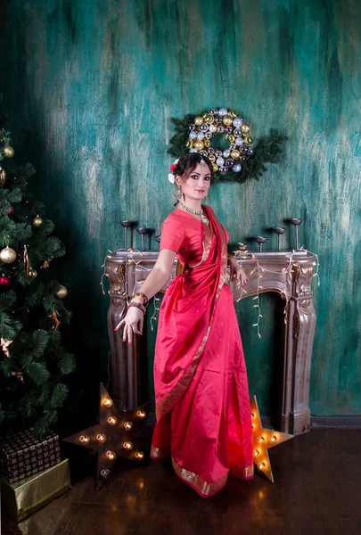 Una niña en un sari escarlata y joyas indias está junto a un árbol de Navidad en el fondo de la chimenea y la pared de color verde oscuro, extendiendo su brazo en un gesto de la danza . —  Fotos de Stock