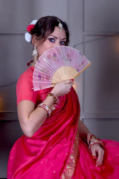 La chica en un sari escarlata y joyas indias se sienta mirando hacia fuera sobre un ventilador sobre un fondo de pared gris —  Fotos de Stock