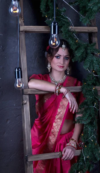 La chica en un sari escarlata y joyas indias asomándose por la escalera de madera y bombillas colgando del techo —  Fotos de Stock