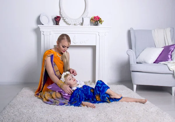 Mujer rubia joven en un brillante saris indio y joyas sentadas en el suelo frente a una chimenea blanca, arrodillando la cabeza de su hija — Foto de Stock