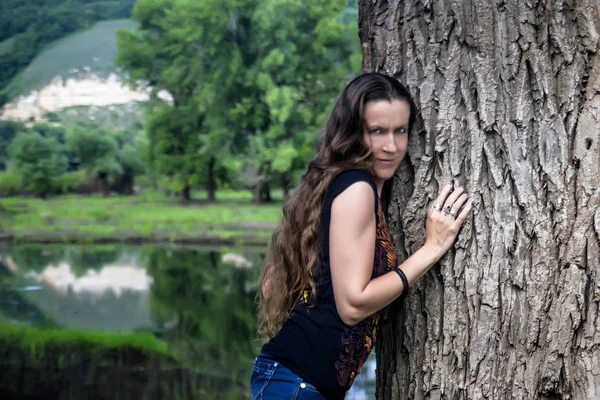 Girl with long golden hair in jeans and black T-shirt pressed her cheek to trunk of a huge tree on the background of forest lake