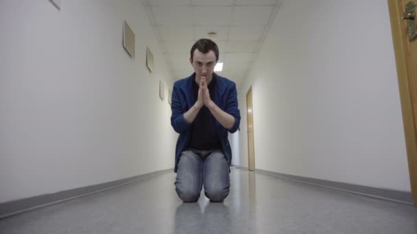Молодой человек молится на коленях в белом коридоре — стоковое видео