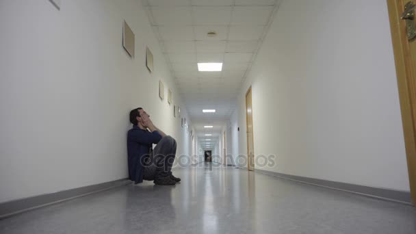 Молодой человек приседает, склоняясь к стене в длинном коридоре и ожидая — стоковое видео