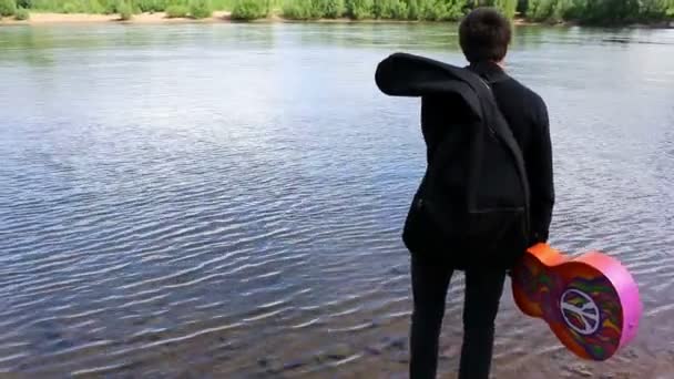 Man walks edge of river with acoustic guitar seeking inspiration for songwriting, music. hippie. — Stock Video