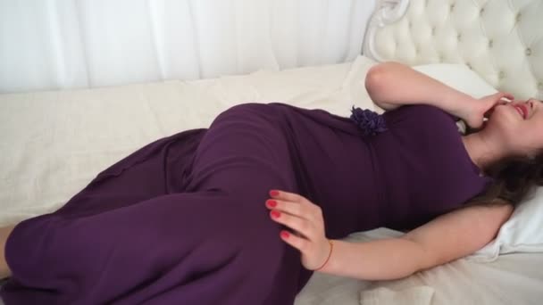Молодая женщина в фиолетовом платье лежит на белой кровати и мечтает — стоковое видео