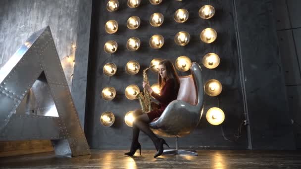 Девушка в платье с саксофоном сидит на поворотном стуле в ретро-комнате рядом с большой буквой А — стоковое видео