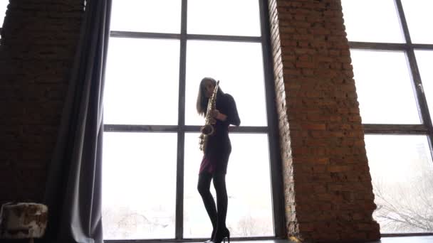 Девушка в коротком платье играет на саксофоне в ретро-комнате возле большого окна — стоковое видео
