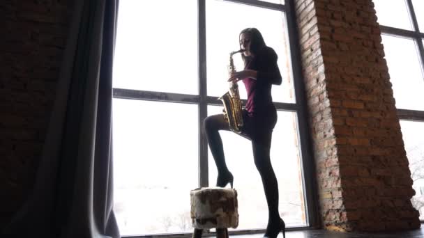Κορίτσι σε σύντομο φόρεμα παίζει σαξόφωνο σε ρετρό χώρο κοντά σε μεγάλο παράθυρο — Αρχείο Βίντεο