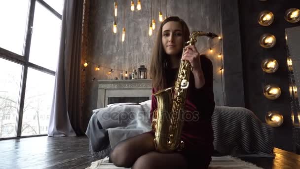 Hezká dívka v šatech s saxofon na sedět na parapetu v retro místnosti u okna — Stock video