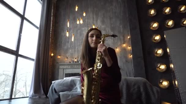 Mooi meisje in jurk met saxofoon op zittend op de vensterbank in retro kamer in de buurt van venster — Stockvideo