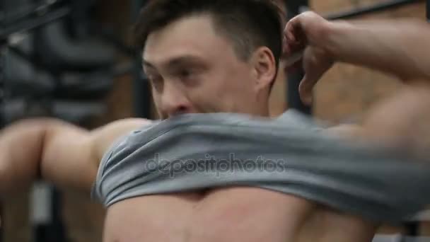 Culturista poniéndose una camiseta gris en el fondo del gimnasio — Vídeos de Stock