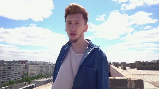 Κοκκινομάλλα νεαρός άνδρας στέκεται στη στέγη σε φόντο του αστικού τοπίου και μπλε ουρανού — Αρχείο Βίντεο