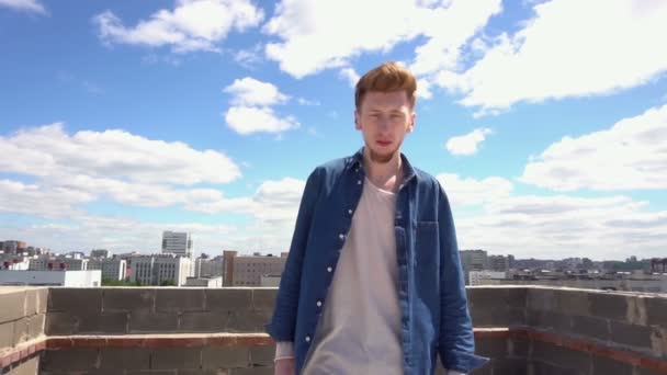 Κοκκινομάλλα νεαρός άνδρας είναι το περπάτημα στην οροφή σε φόντο από το αστικό τοπίο και το γαλάζιο του ουρανού — Αρχείο Βίντεο