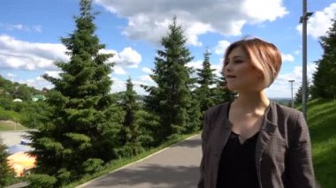 Ağır çekim mutlu gülümseyen genç kadın ceket çevresinde güneşli parkta yaz gününde yürüyüş.