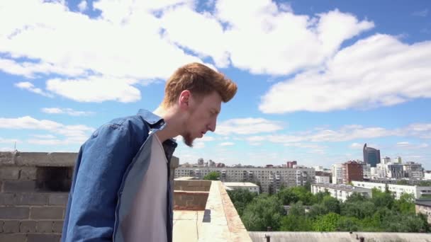 Рыжий молодой человек идет по крыше на фоне городского пейзажа и голубого неба — стоковое видео