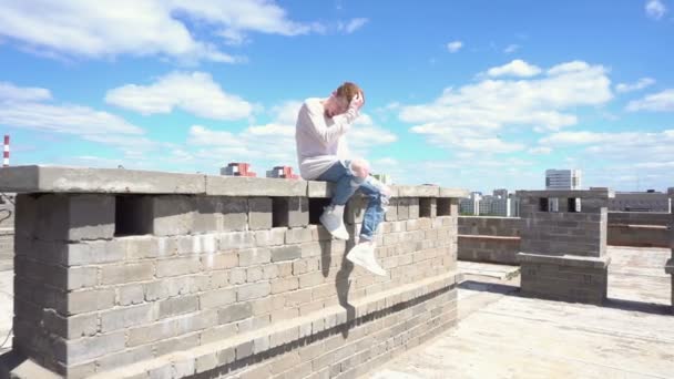 Joven pelirrojo con una sudadera blanca se sienta en una pared de ladrillo en el techo — Vídeos de Stock