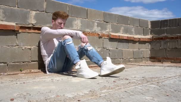 En ung rödhårig man sitter på golvet taket i en varm solig dag — Stockvideo