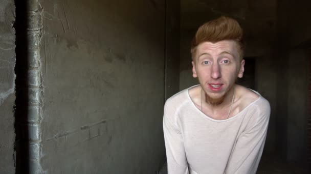 Молодой человек с рыжей бородой танцует и улыбается в камеру в сером бетонном коридоре или подвале — стоковое видео