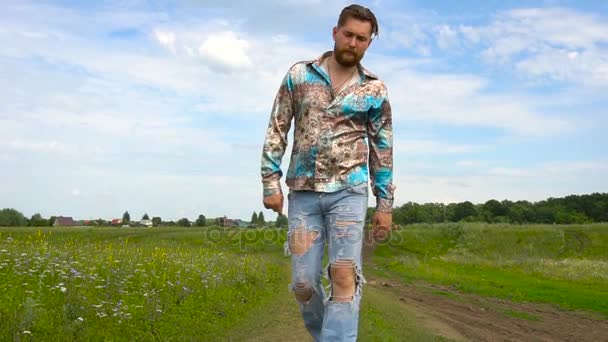 En man med ett skägg i rippade jeans promenader längs fältet på en sommardag — Stockvideo