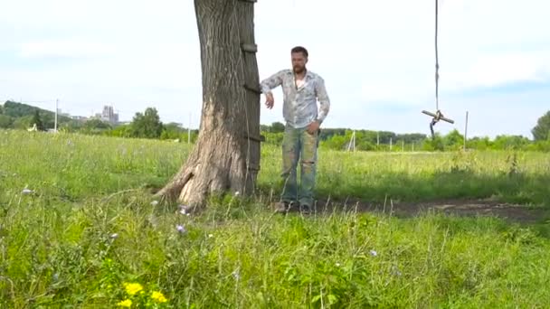 Красивий бородатий чоловік спирається на дерево в полі — стокове відео