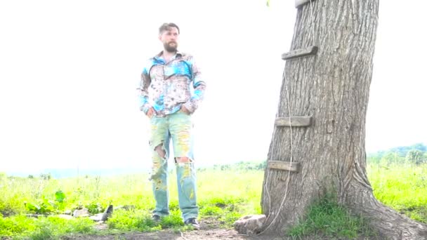 Hombre barbudo guapo se apoya en un árbol en un campo — Vídeo de stock