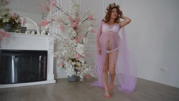 Grossesse belle jeune femme en lilas peignoir et couronne de tête florale marche sur la chambre de luxe — Video