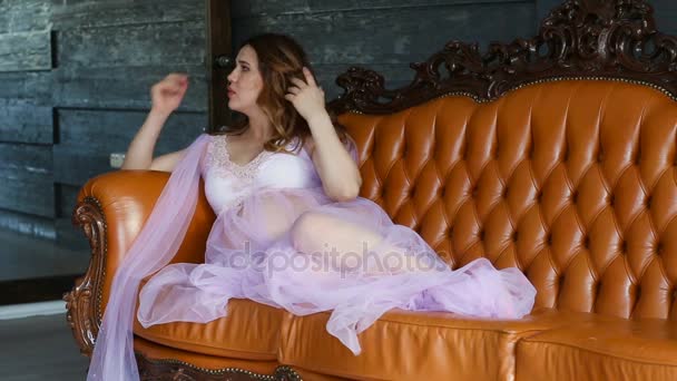 Joven y hermosa mujer embarazada endereza su peignoir mientras está sentado en el sofá — Vídeos de Stock