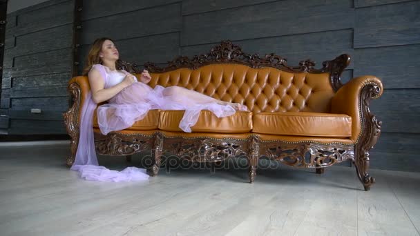 Красивая очаровательная беременная женщина с каштановыми волосами сидит на диване в лиловом винтажном платье — стоковое видео
