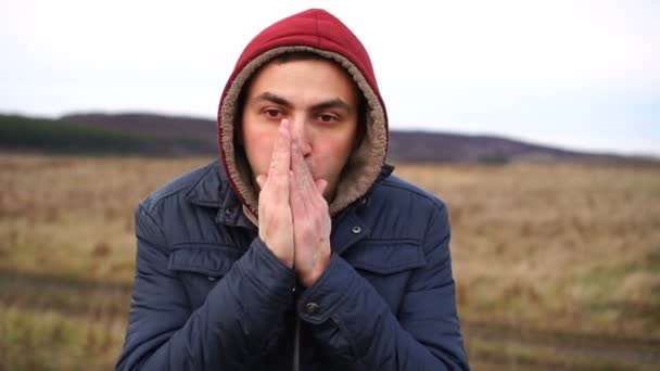 Παγωμένος άνθρωπος στέκεται σε ένα φθινοπωρινό χωράφι προσπαθώντας να ζεσταθεί. Τα μάτια κλειστά και φυσώντας ζεστό αέρα στα χέρια του. — Αρχείο Βίντεο