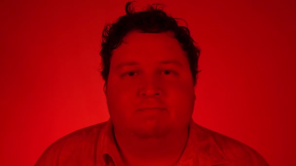 Portrait tourné avec un éclairage rouge : Gros homme debout isolé sur fond rouge et cligner des yeux tout en regardant la caméra — Video