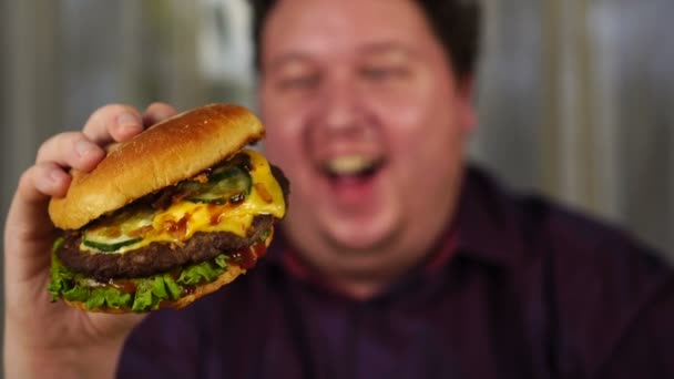 Молодой человек держит гамбургер. Толстяк ест фаст-фуд. Бургер не помогает с едой. Очень голодный парень . — стоковое видео