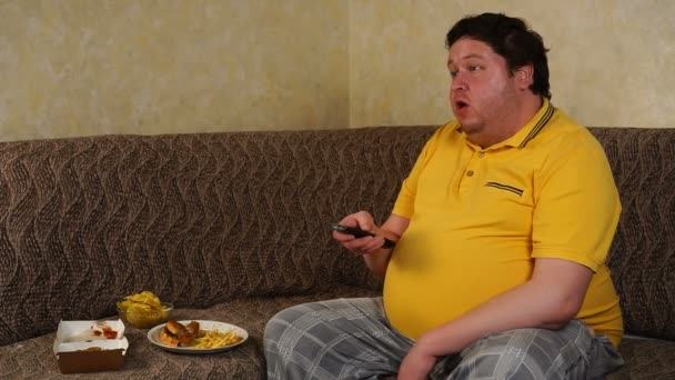 Tlustý muž sedí doma a dívá se na televizi a jí fast food — Stock video