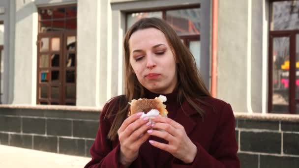 Chica atractiva en abrigo come hamburguesa en la calle — Vídeos de Stock