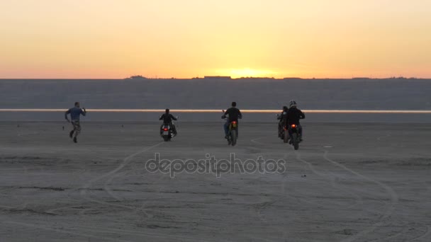 Bikers ride into sunset on lake, river, sea, bay. Man flees, falls and tumbles. — Stock Video