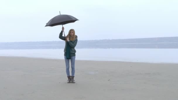 Hübsches junges Mädchen, das Regenschirm dreht und Spaß am nebligen Strand hat — Stockvideo