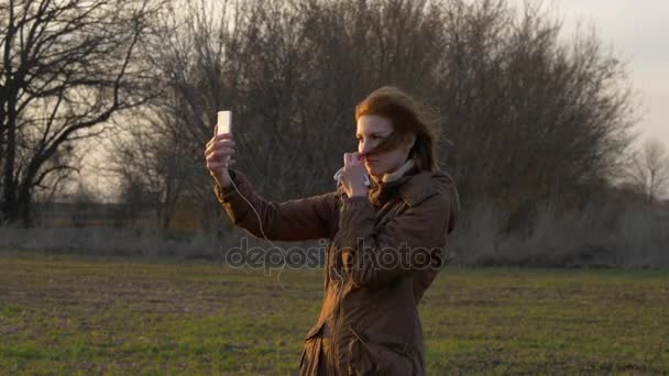 Μια κοκκινομάλλα κοπέλα κάνει μια selfie με μια κάμερα για smartphone κοντά σε δέντρα. — Αρχείο Βίντεο