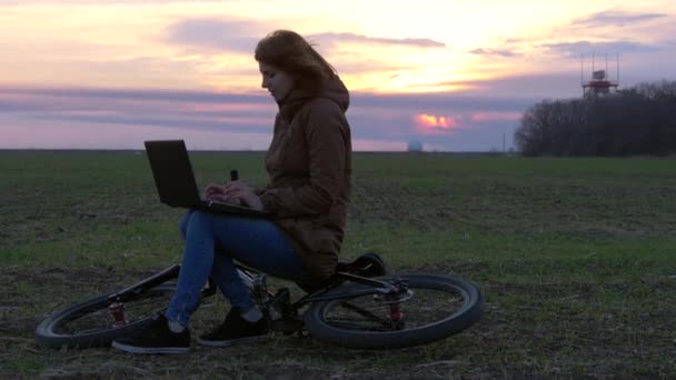 Рыжая девушка использует ноутбук возле радара в поле во время захода солнца на велосипеде — стоковое видео