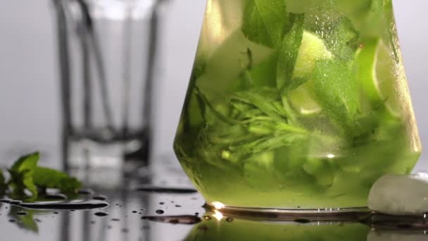 Koud vers mojito limonade met munt en limoen in een kruik. Limonade vult een glas met een buis in de achtergrond. Slow motion. — Stockvideo