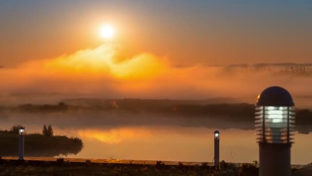 Flusspier mit brennenden Laternen gegen den Zeitraffer des sich bewegenden Nebels auf dem Fluss und die atemberaubende Morgendämmerung mit einer massiven hellen Sonne, die hinter dem Horizont aufgeht. — Stockvideo