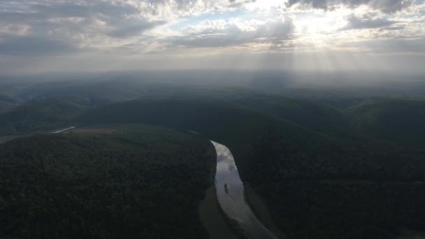 Tiro aéreo de montanhas cobertas com floresta de coníferas com rio e raios de sol através das nuvens . — Vídeo de Stock