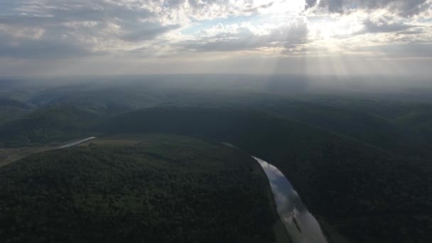 Tiro aéreo de montanhas cobertas com floresta de coníferas com rio e raios de sol através das nuvens . — Vídeo de Stock