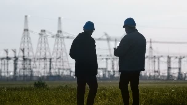 Wide shot of two engineers in hard hat discussing something talking on a smartphone during an energy substation inspection. — Stock Video