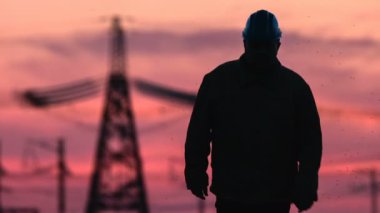 Gözetmen silueti sert şapkalı bir enerji Trafo Merkezi muayene sırasında kırmızı batımında güç hatları boyunca gidiş orta atış.