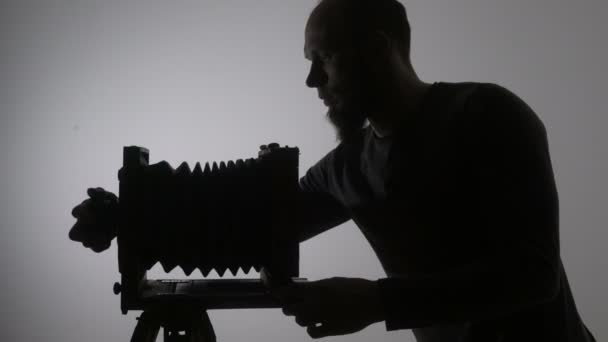 Tiro médio de fotógrafo barbudo ajustando câmera de foto de madeira vintage velho e tirar foto contra a luz no fundo preto . — Vídeo de Stock
