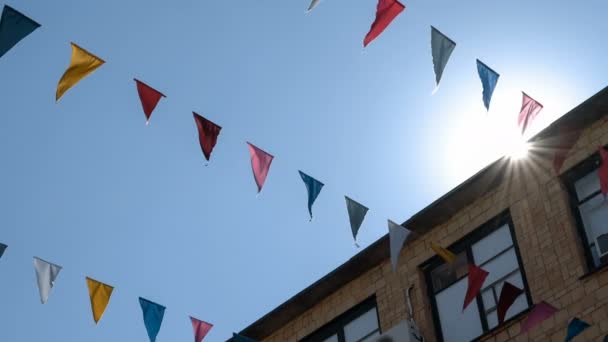 Guirnalda de banderas multicolores de forma triangular se balancea en el cielo azul contra los rayos del sol . — Vídeo de stock