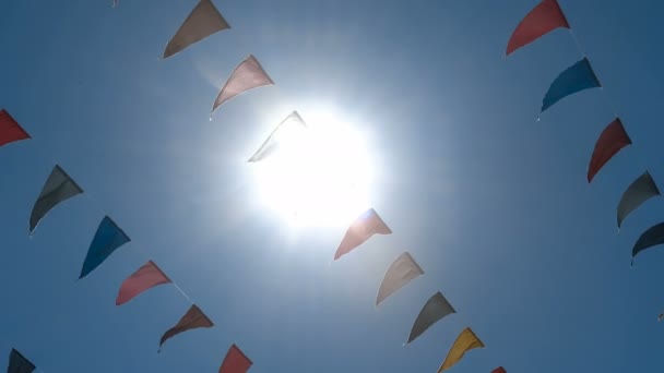 Garland of multi colored flags of triangular shape sways in the blue sky against sun rays. — Stock Video